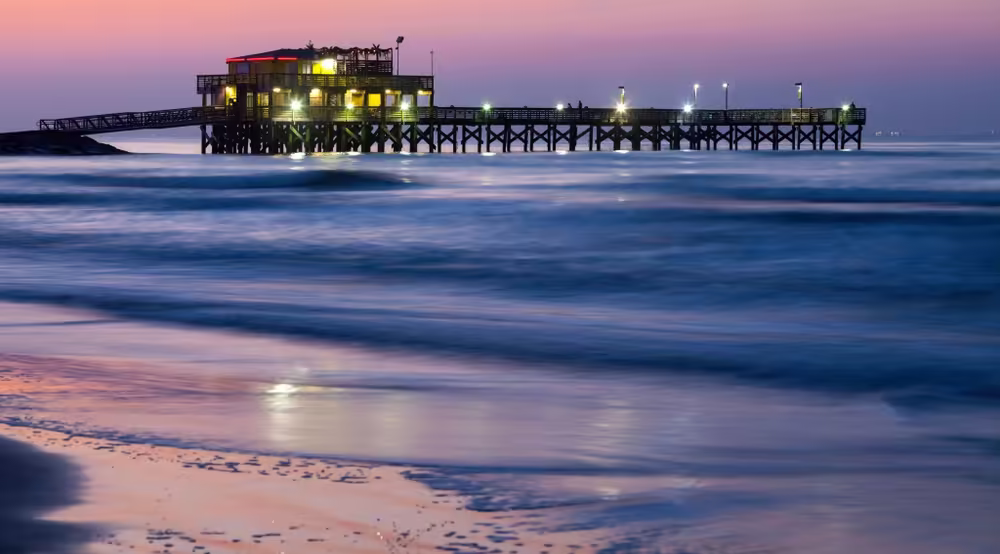 Galveston Wooden fishing pier extending over the ocean at sunset, with soft waves and illuminated structure above the water